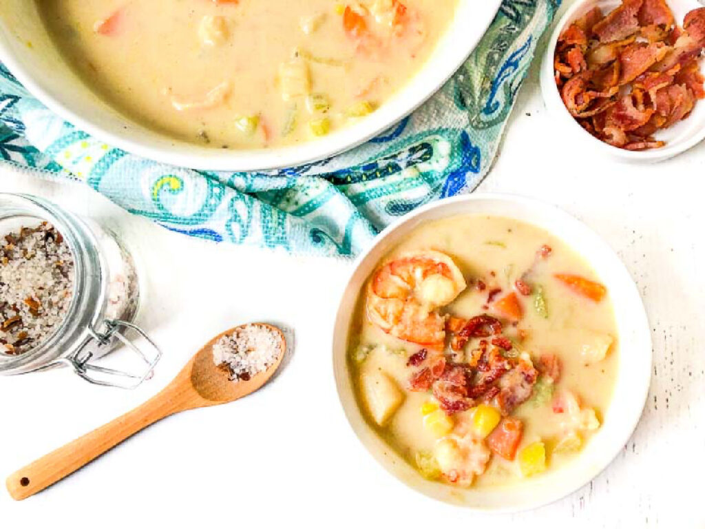 Aerial view of a bowl of the seafood chowder with a bowl of bacon bits and bigger bowl in the background.