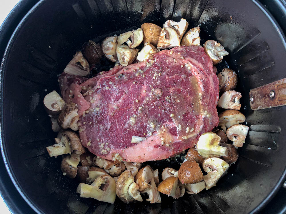 air fryer basket with the uncooked steak and mushrooms covered in garlic butter
