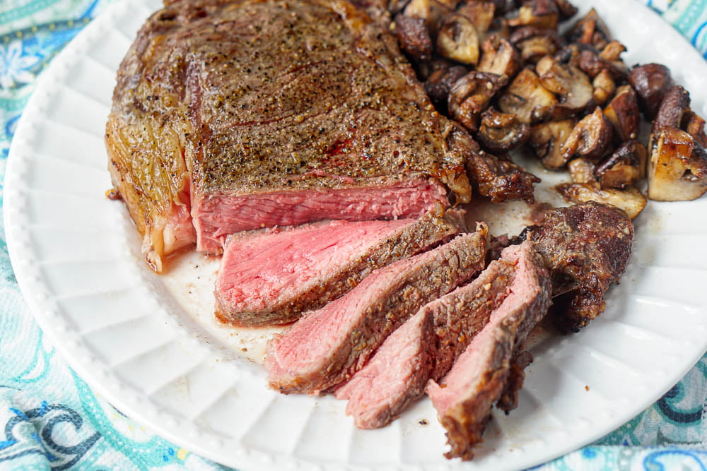 closeup of a rare cooked keto ribeye steak sliced and garlic buttered mushrooms along side all on a white plate