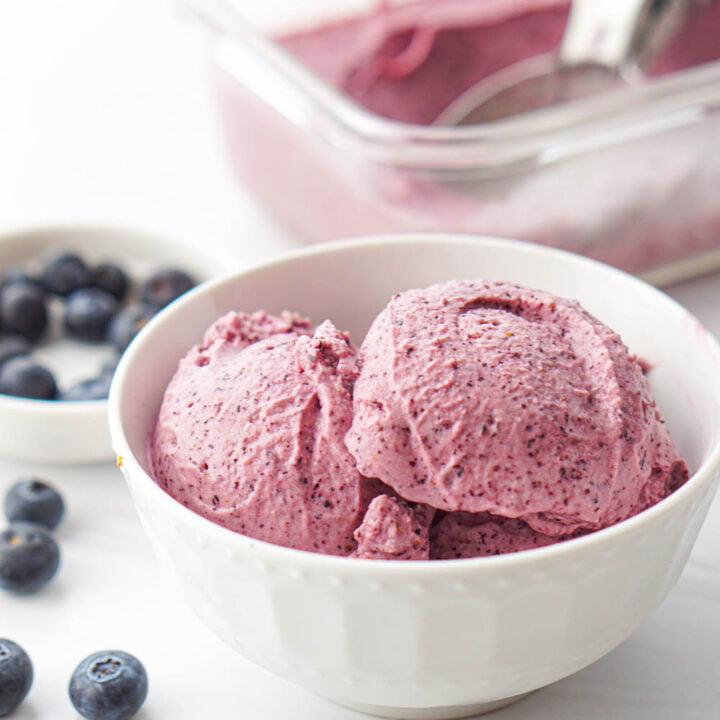closeup of a white bowl with keto blueberry ice cream with scatter blueberries in the background