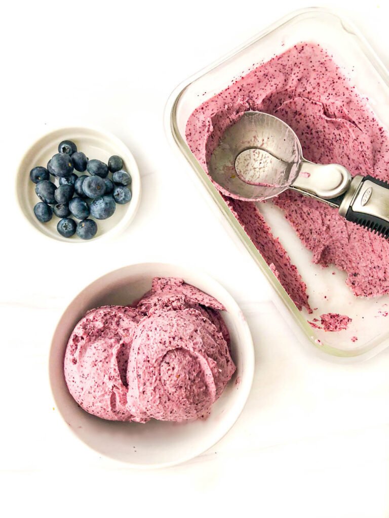 white bowl with sugar free blueberry ice cream and a glass container with the ice cream and a scoop