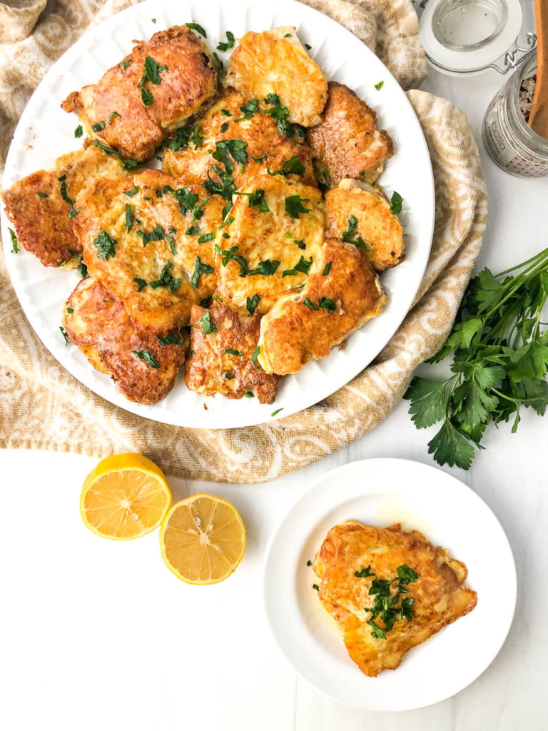 aerial view of a large and small white plate with pieces of keto tilapia and some fresh parsley, lemons and a jar of salt
