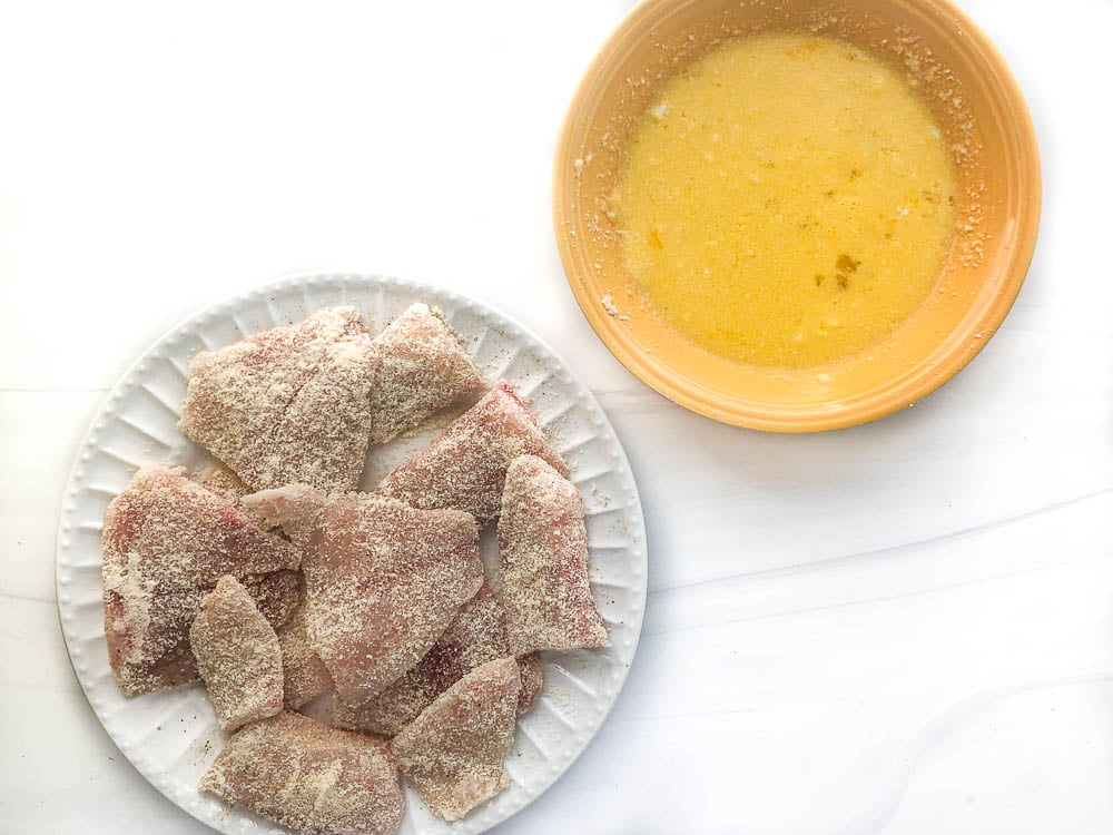 a white plate with pieces of raw tilapia covered in almond flour and a yellow bowl with egg batter