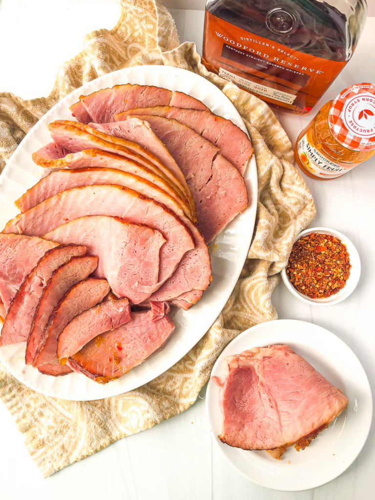 aerial view of a platter of sliced ham and a bottle of bourbon, apricot jam and hot pepper flakes
