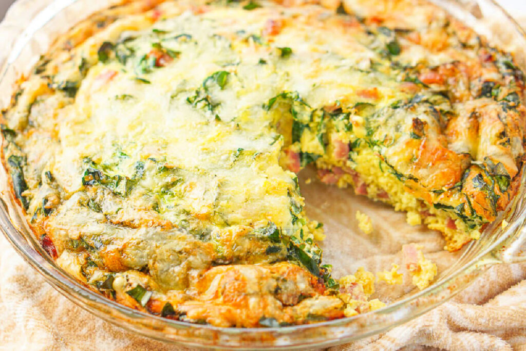 Closeup of the pie plate with a slice of the keto quiche missing so you an see the fluffy eggs, ham and spinach.