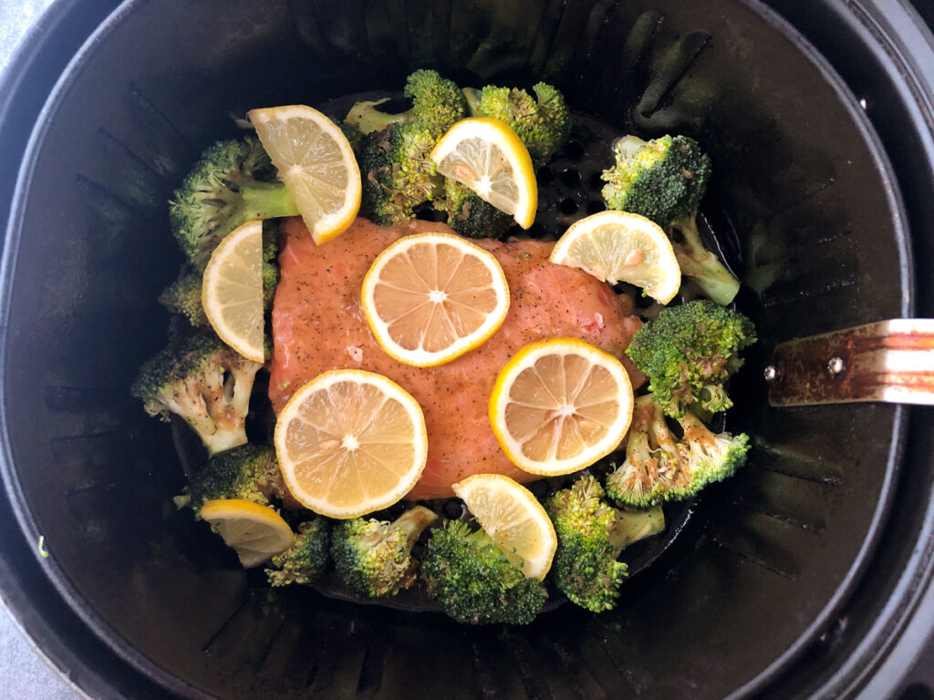 air fryer basket with a raw salmon and broccoli with lemon pepper sauce poured over and fresh lemon slices ready to bake