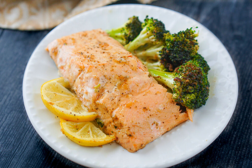 white plate with a piece of cooked salmon with lemon sauce and roasted broccoli and lemon slices