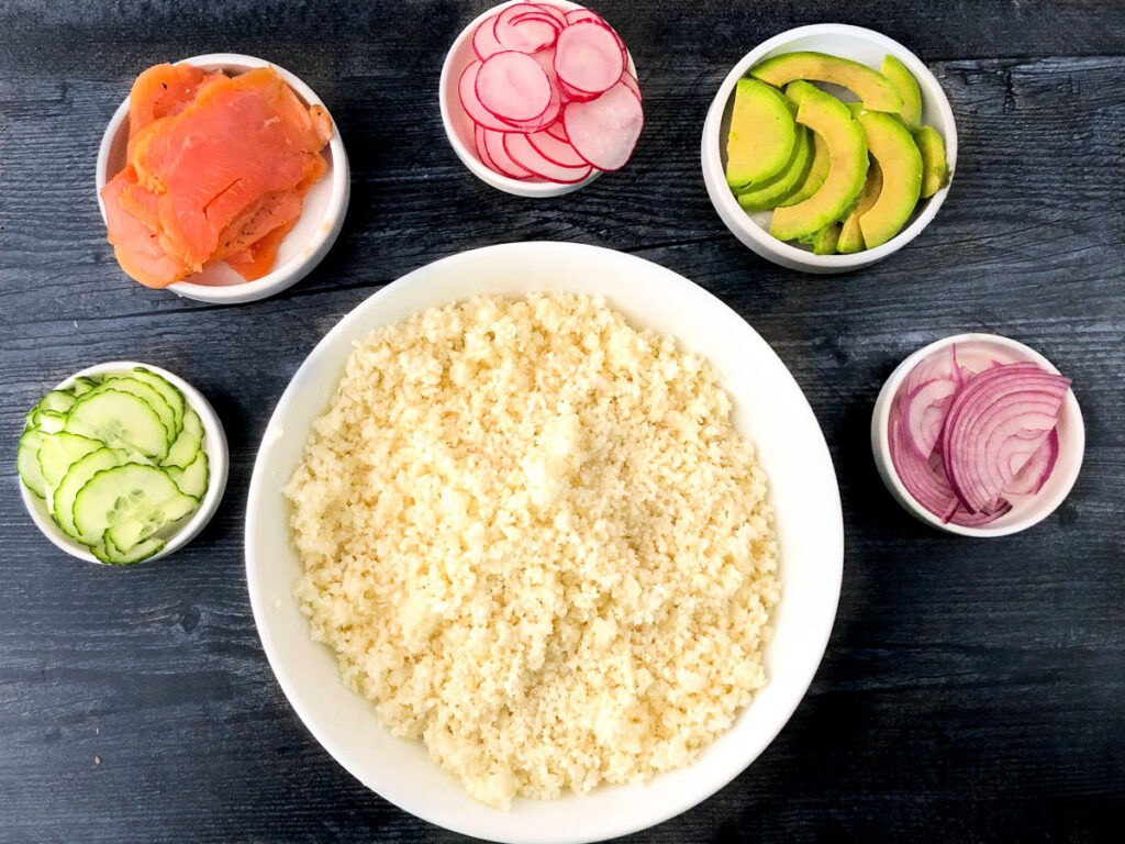 white bowl with cauliflower rice and smaller bowls with smoked salmon, cucumber, radishes, avocado and red onion