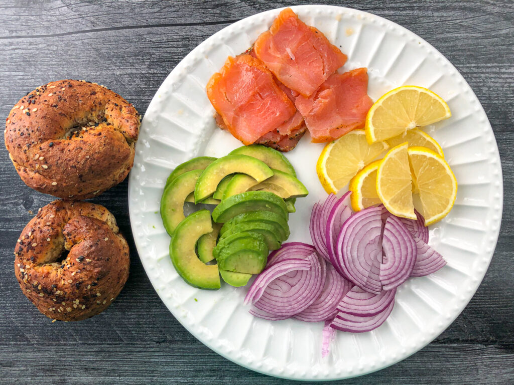 ingredients for the bagel sandwich - everything bagels, and white place with smoked salmon, avocado, lemon, and onions