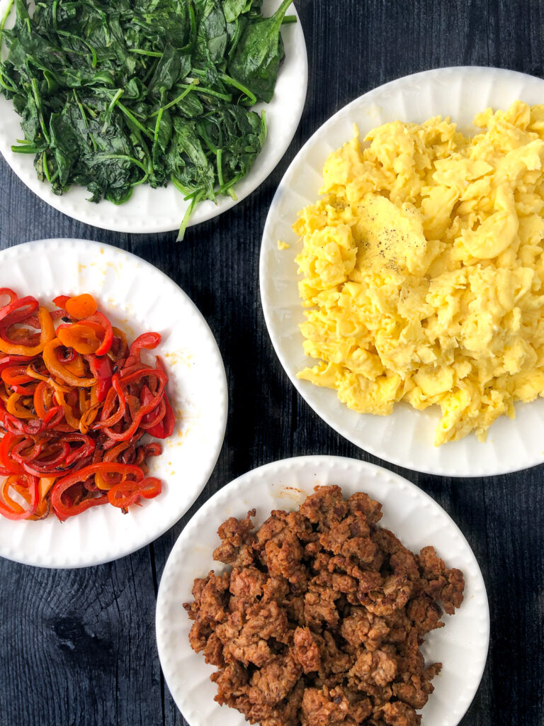 white plates with ingredients to make breakfast bowls - scrambled eggs, sautéed spinach, sautéed peppers and turkey sausage