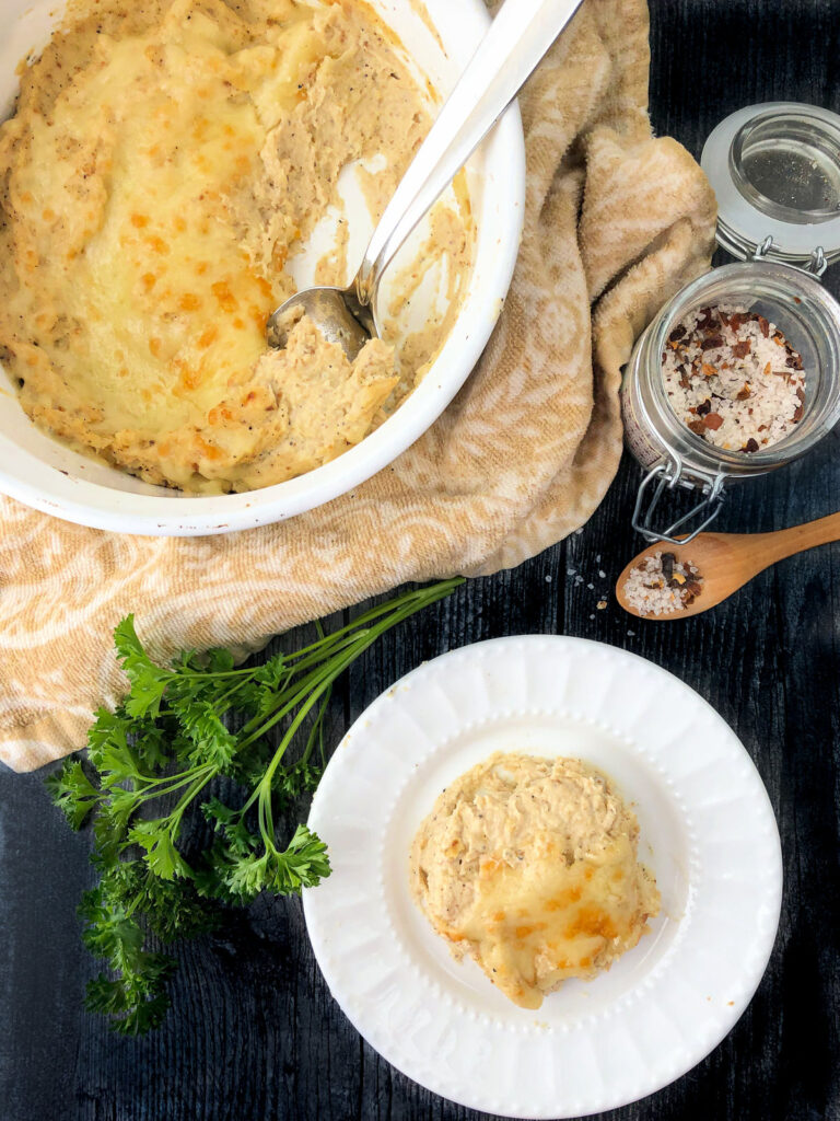 aerial view of a baking dish and white plate with mashed cheesy cauliflower and parsley sprigs