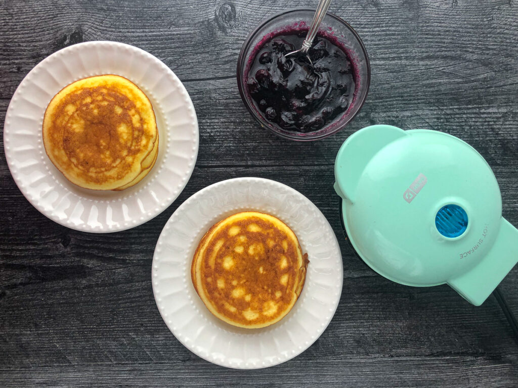 aerial view of low carb pancake on white plates and a bowl of blueberry sauce and a mini dash griddle