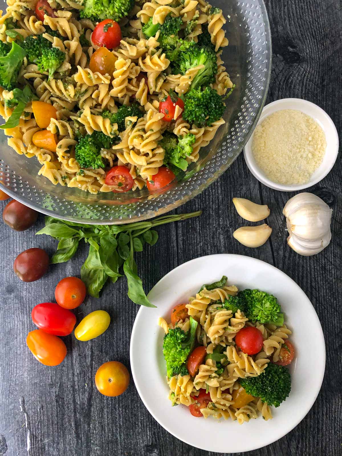 glass bowl and white plate with broccoli pesto pasta salad and fresh  basil and garlic with heirloom tomatoes