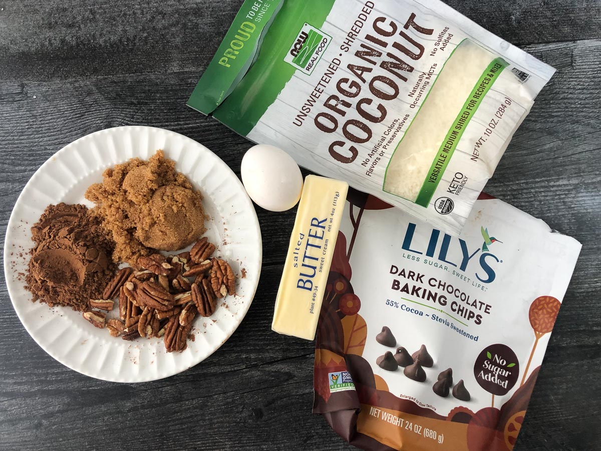 keto coconut cookie ingredients - brown sweetener, cocoa powder, pecans, coconut, butter and Lily's chocolate chips and an egg