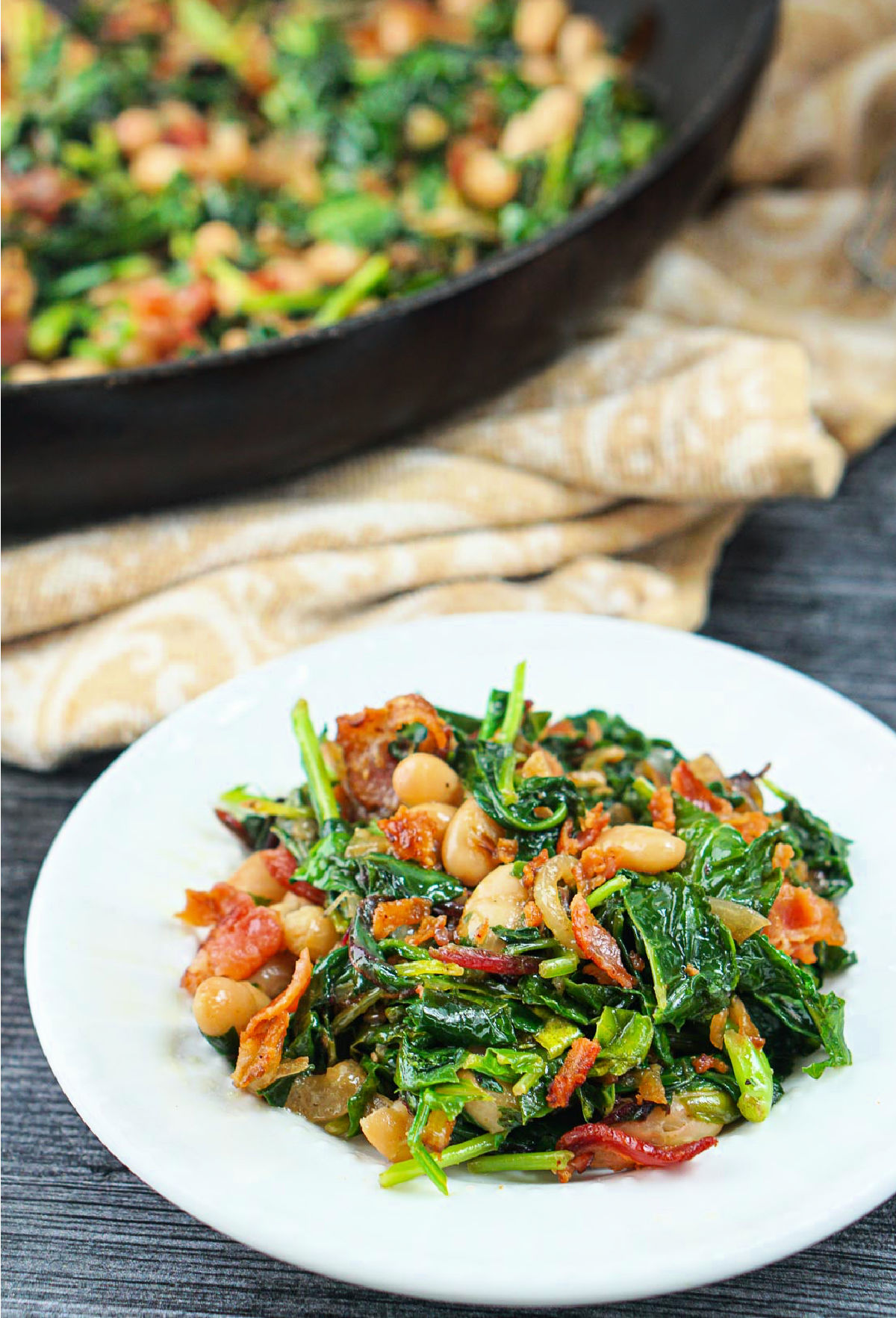 White plate with a serving of white beans and greens with bacon and pan of it in the background.