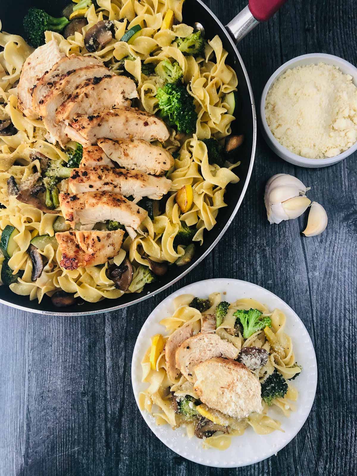 aerial view of a skillet with chicken and pasta with garlic vegetables and a plate with a serving and parm cheese