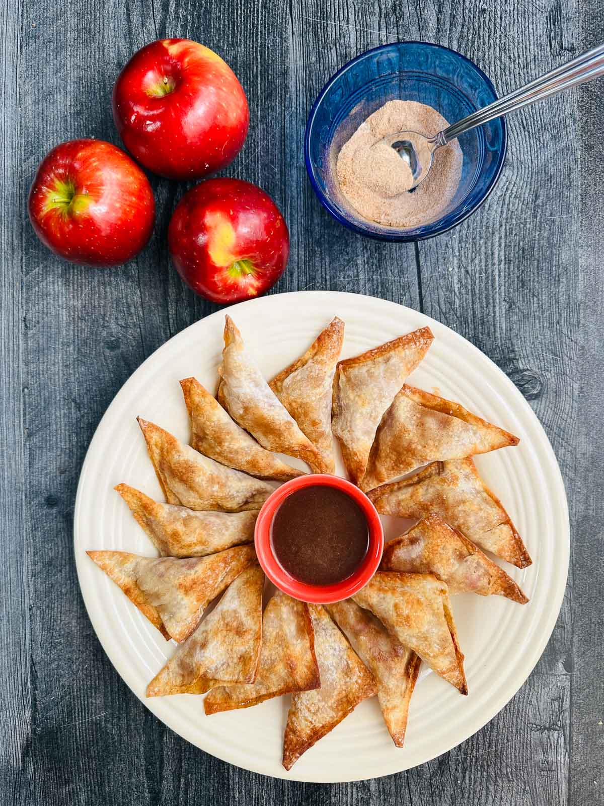Aerial view of a plate of the apple wontons with a a bowl of cinnamon brown sugar sauce and fresh apples and cinnamon sugar.