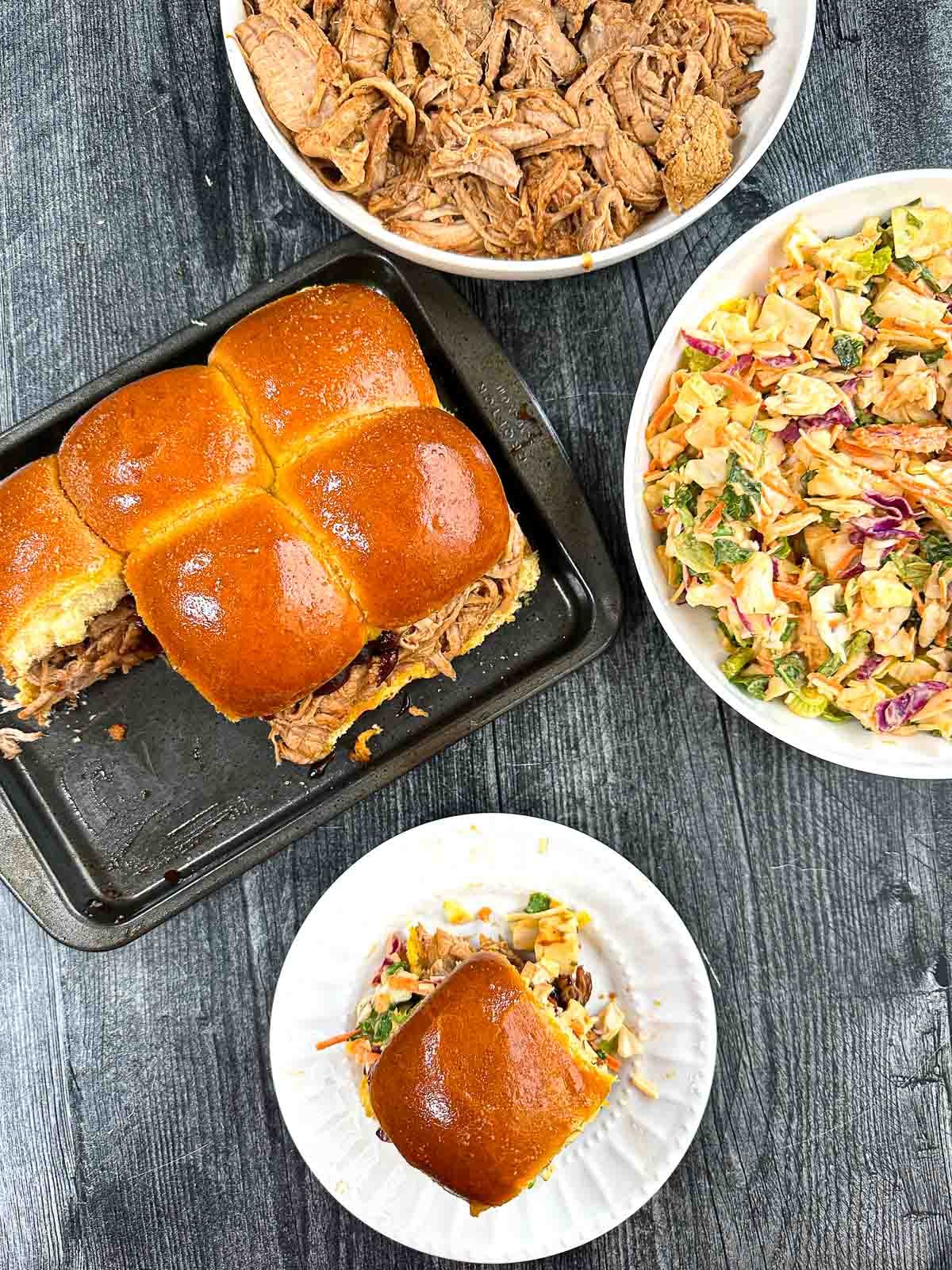 Aerial view of the bbq sliders on a baking tray and white plate with a bowl of pulled pork and a bowl of slaw.