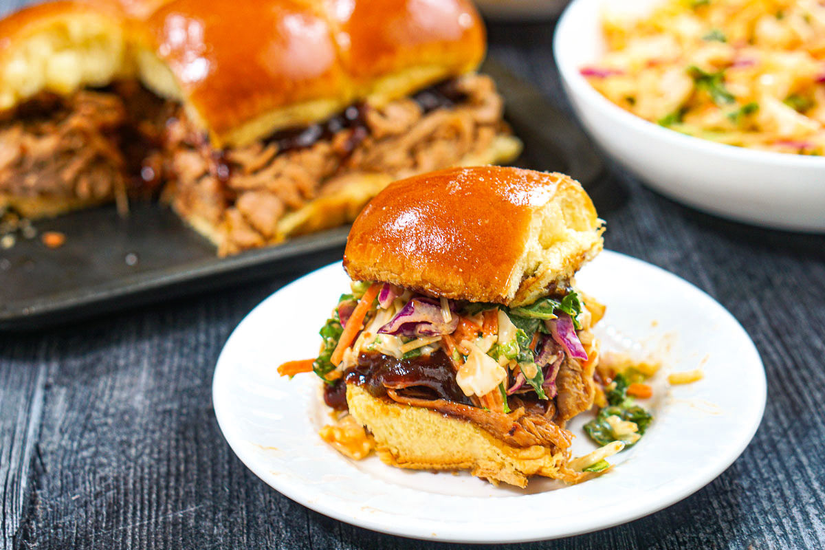 Closeup of a white plate with a bbq slider with a tray of them in the background.