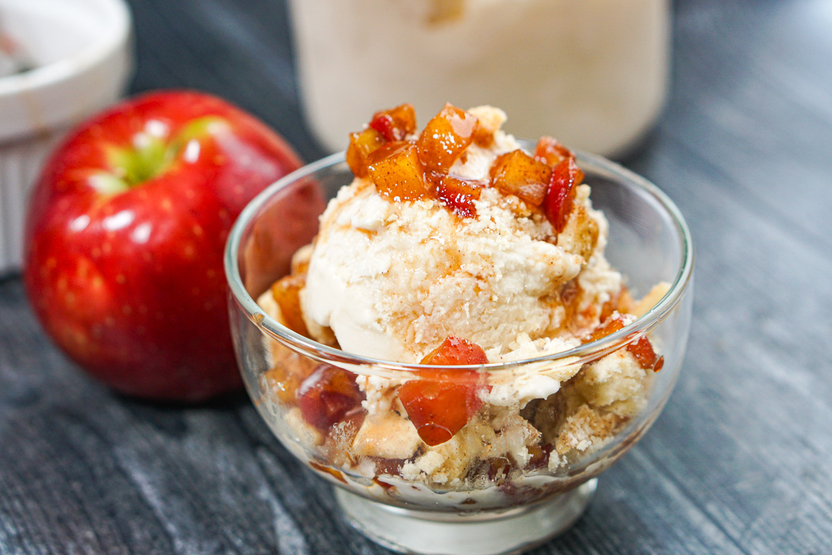 Closeup of a dessert cup with low carb Ninja Creami apple pie ice cream with diced cinnamon apples and pie crust crumbles with apple in background and container of ice cream.