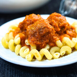 White plate with pasta and meatballs with veggies covered in sauce.