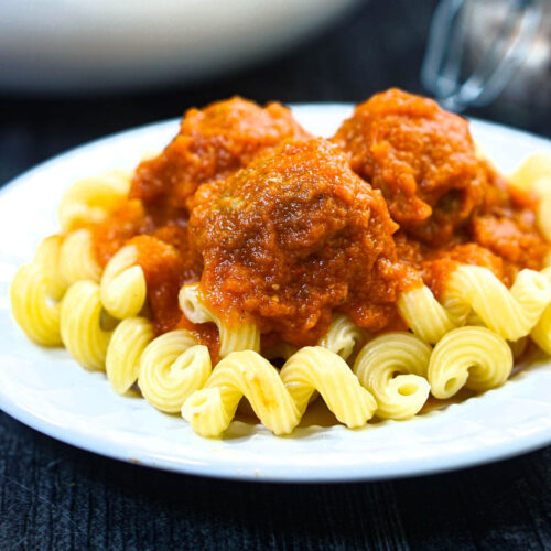 White plate with pasta and meatballs with veggies covered in sauce.