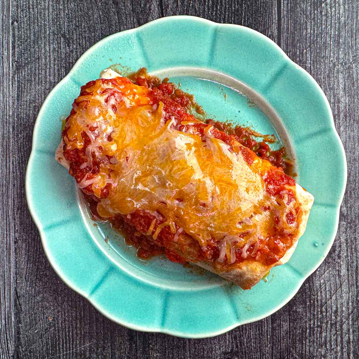 Aqua plate with cooked chicken cheese meal prep burritos with salsa and cheese on top.