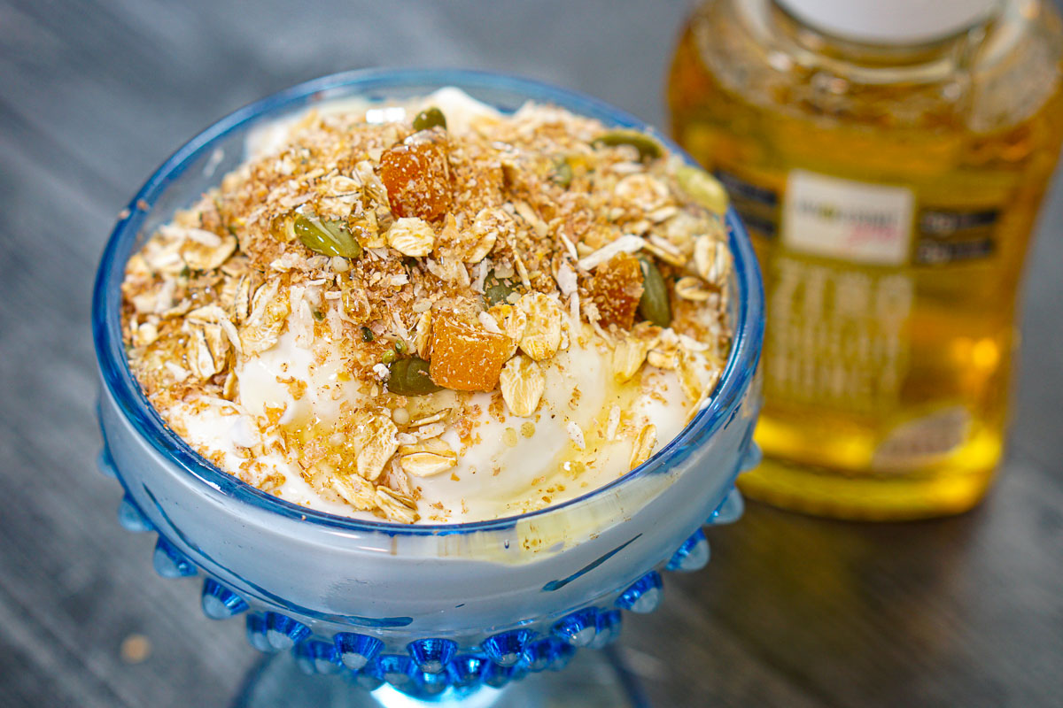 Closeup of a blue dessert dish with yogurt and muesli on top and low carb honey in back.