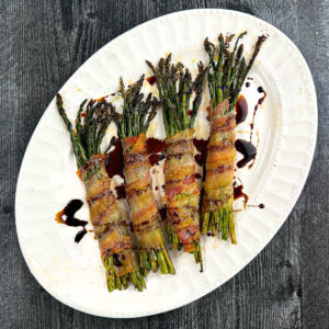 White platter with bundles of bacon wrapped asparagus and balsamic glaze.