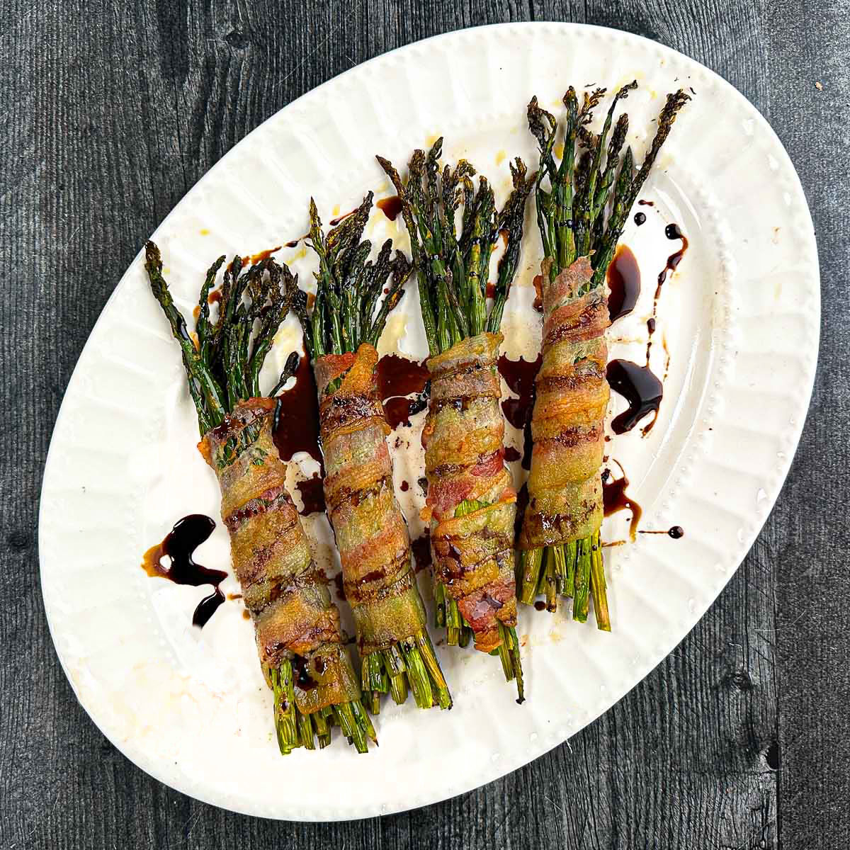 White platter with bundles of bacon wrapped asparagus and balsamic glaze.