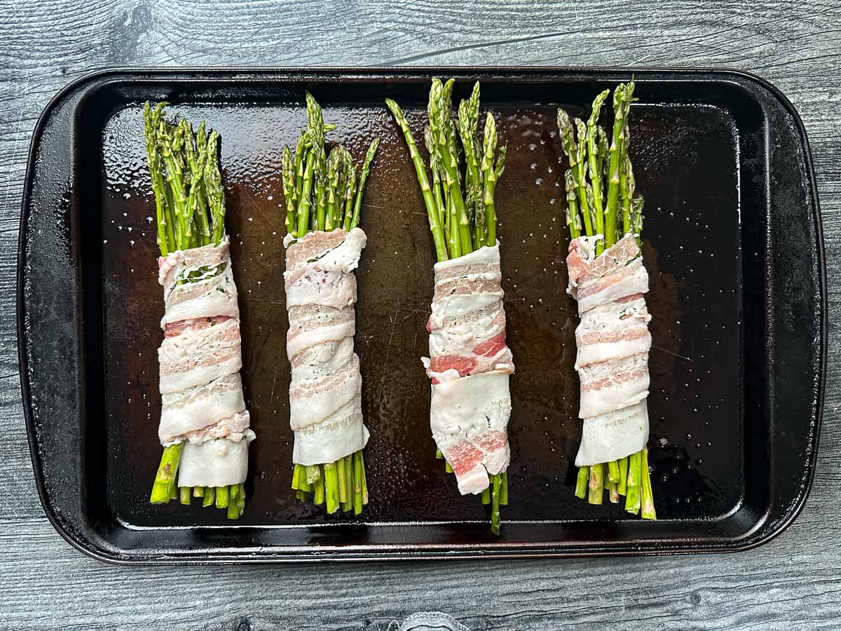 Baking sheet with bundles of asparagus spears wrapped in slices of raw bacon.