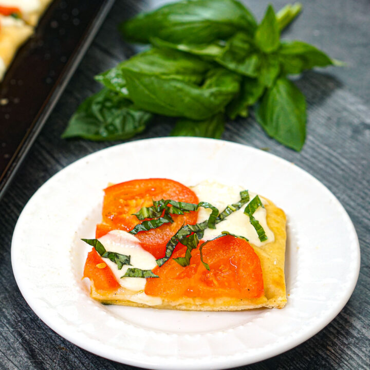 White plate with a piece caprese flatbread with a a sprig of basil in the background.