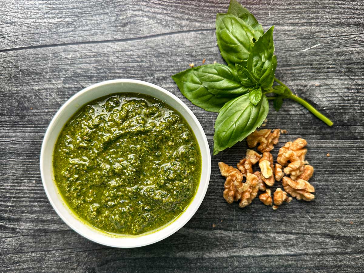 White bowl with the finished pesto and fresh basil and walnuts beside it.